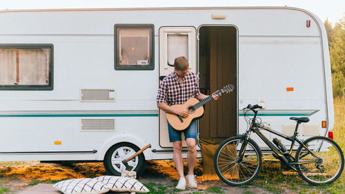 A man with a guitar stands near a trailer at sunset