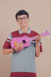 Portrait of smiling young man playing guitar while standing against brown background