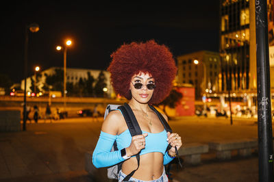Confident female with afro hairstyle and trendy outfit with backpack looking at camera while standing on street with buildings in evening time