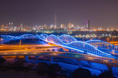 Illuminated bridge over river by buildings against sky at night