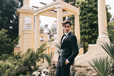 A beautiful young man, the groom in an elegant wedding suit, stands posing in the city's old park