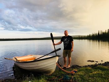 Portrait of mature man with boat standing at lakeshore against cloudy sky during sunset