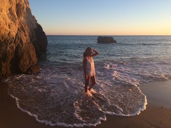 Woman walking on shore at beach against sky during sunset