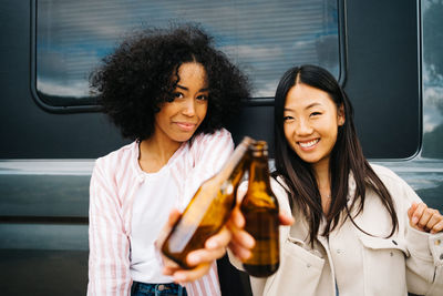 Happy young multiethnic female friends clinking bottles of beer while having fun and enjoying summer holidays together near camper van parked in nature