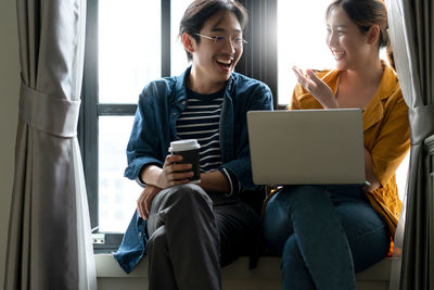 Smiling young woman using phone while sitting on window