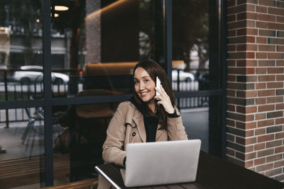 Portrait of smiling young woman using phone while sitting on table