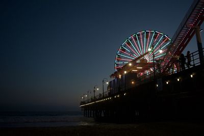 Low angle view of illuminated ferris wheel on pier by sea at night