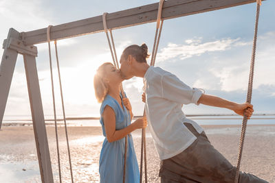 
close-up of a man and a woman riding on a swing against the rising sun on the seashore