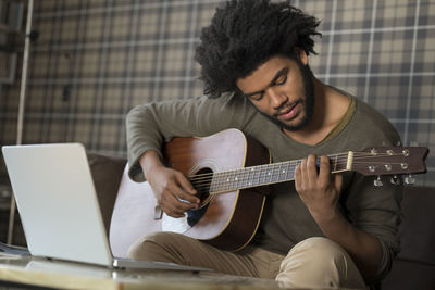 Man sitting in living room on sofa playing guitar in front of laptop