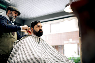 Male barber with comb brushing hair of ethnic hispanic client while making hairstyle in modern barbershop