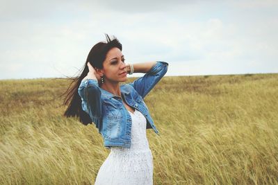 Beautiful woman with hand in hair standing on grassy field against sky