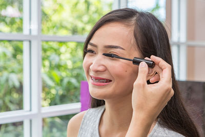 Smiling woman applying mascara against window at home