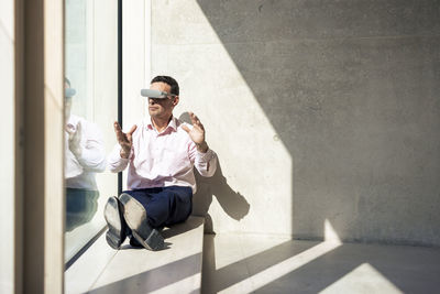 Businessman with vr glasses gesturing sitting on window sill in office