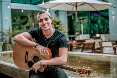 Portrait of happy young man holding guitar while sitting on railing