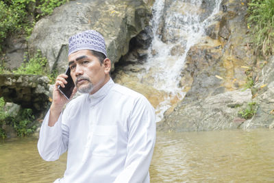 Thoughtful man talking on phone while sitting against waterfall