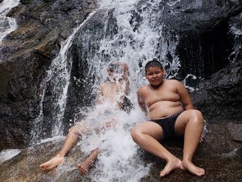 Full length of shirtless young man with boy sitting on rock under waterfall