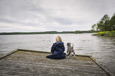 Rear view of senior woman and dog on pier at lake against cloudy sky