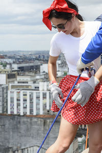 Woman wearing hero costume being prepared by a man to descend a tall rappel building. 