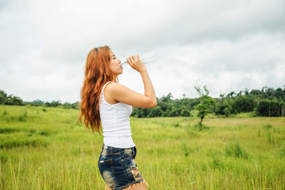 Woman drinking water while standing by plants against sky