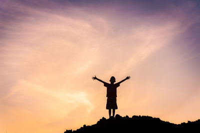 Silhouette boy with arms raised standing against sky during sunset