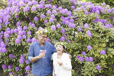 Cheerful senior couple wearing tiaras standing with drinks against flowering plants