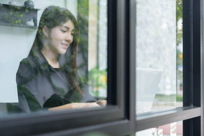 Young woman using laptop in office seen through window