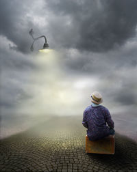 Man with suitcase sitting on road against cloudy sky