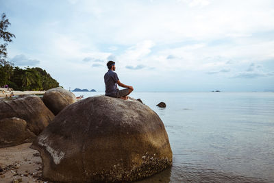 Man sitting on rock looking at sea against sky