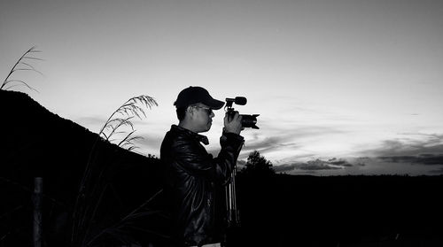 Side view of man photographing against sky during sunset