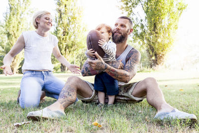 Father holding son and ball by woman while sitting on grass against trees