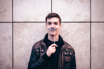 Portrait of young man holding mustache prop while standing against wall