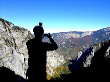 Silhouette mid adult man photographing while standing on mountain against clear blue sky