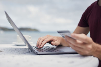 Man sitting on beach against sea and using credit card for online reservation or shopping.