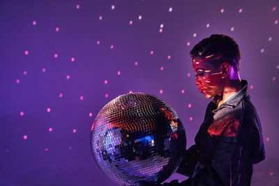 Side view of trendy young african american female with makeup and glitter ball in violet light