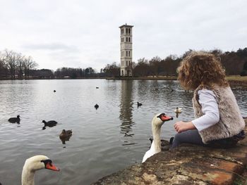 Side view of girl playing with swans while sitting by lake