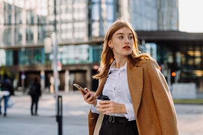 Elegant female entrepreneur reading financial news on phone while passing crosswalk in megalopolis