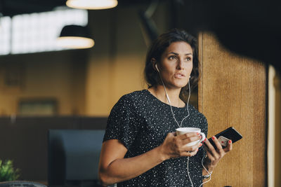 Female executive holding coffee cup while talking with earphones during phone call in office