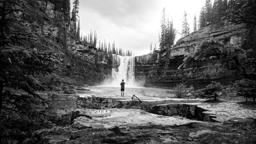 Rear view of person standing on rock against waterfall and sky