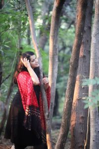 Side view of young woman standing by tree trunk in forest