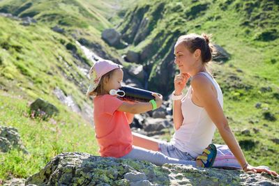 Mother looking at daughter drinking water while sitting on mountain