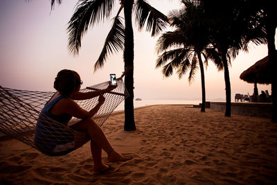 Woman photographing while sitting on hammock at beach during sunset