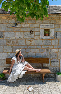 Beautiful happy young woman wearing white dress, sitting on bench in front of old stone house