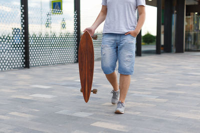 Close up street picture of man in denim shorts and grey sneakers walking with a longboard in hand.