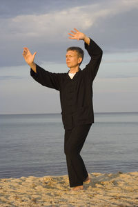 Full length of mature man practicing tai chi on sea shore at beach against cloudy sky