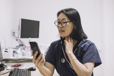 Female otolaryngologist explaining while touching throat during online consultation through smart phone in medical clini