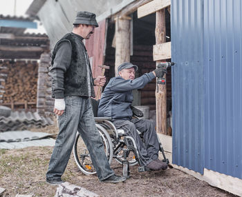 Happy elderly man in casual clothes in a wheelchair is working  in his yard. a friend helps him.