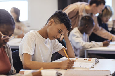 Bored male student writing in book while sitting by female friend at desk