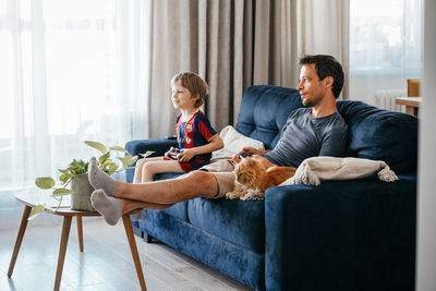 Cheerful father and son playing video games at home sitting on couch with ginger cat