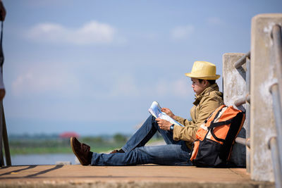 Hiker reading map while sitting on pier against sky