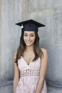 Portrait of young woman smiling while wearing mortarboard against wall